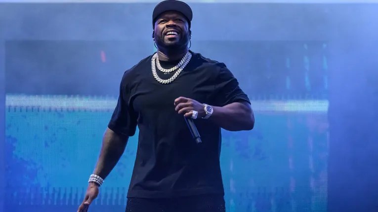 50 Cent on stage in a black tee and heavy chains, gripping a microphone