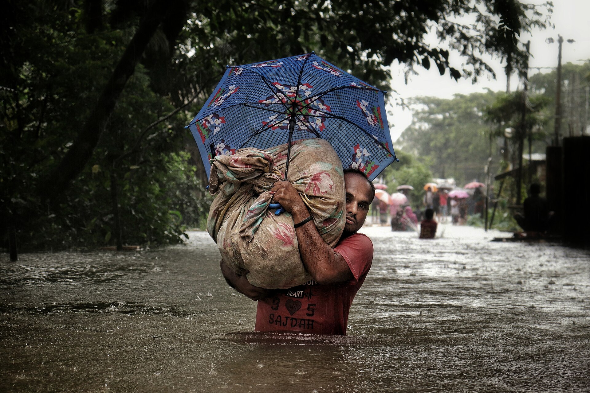 A man wades through floodwater in Sylhet, carrying his belongings in a bundle under an umbrella.