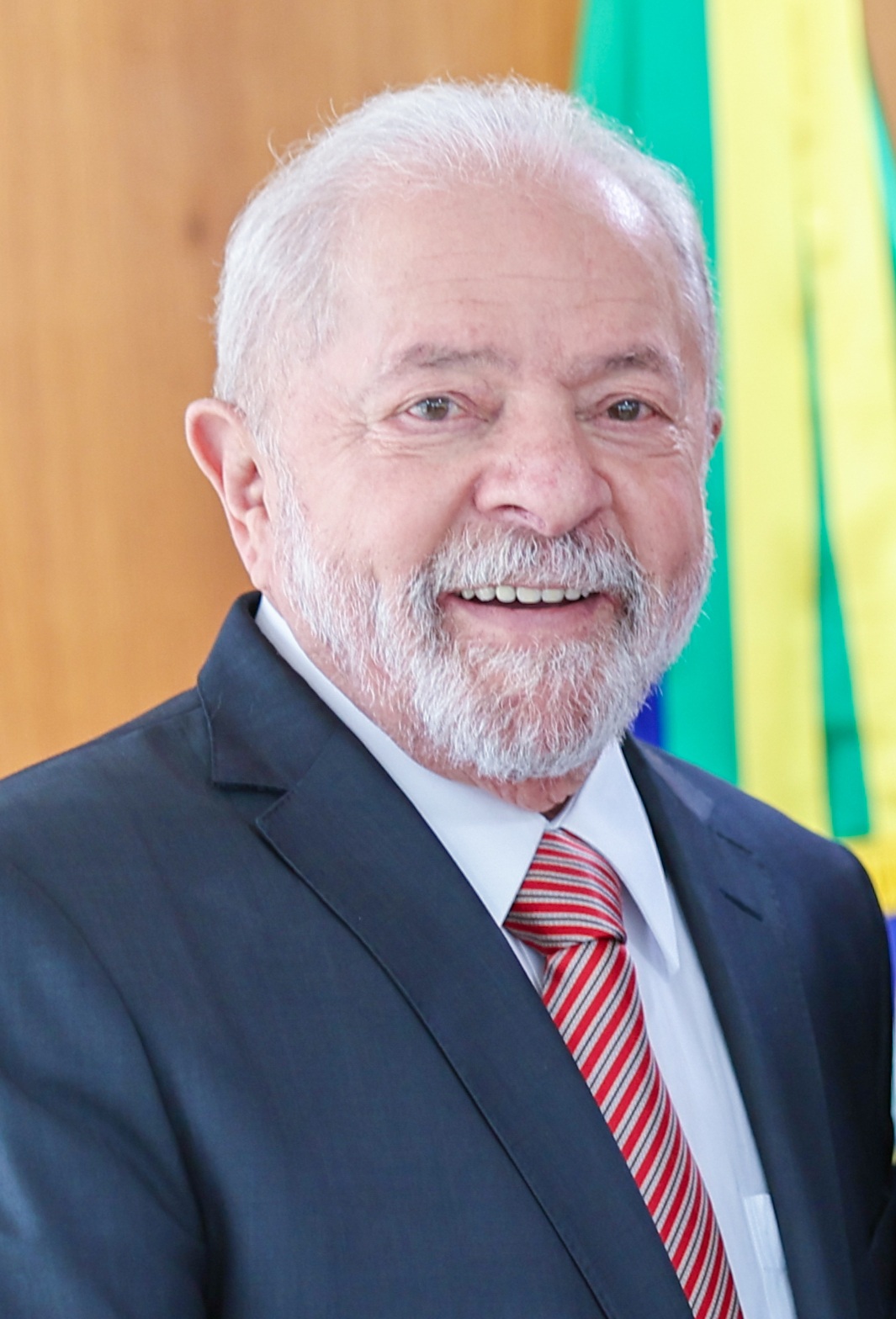 Brazilian President Luiz Inácio Lula da Silva smiling in a dark suit and red striped tie, with the Brazilian flag behind him