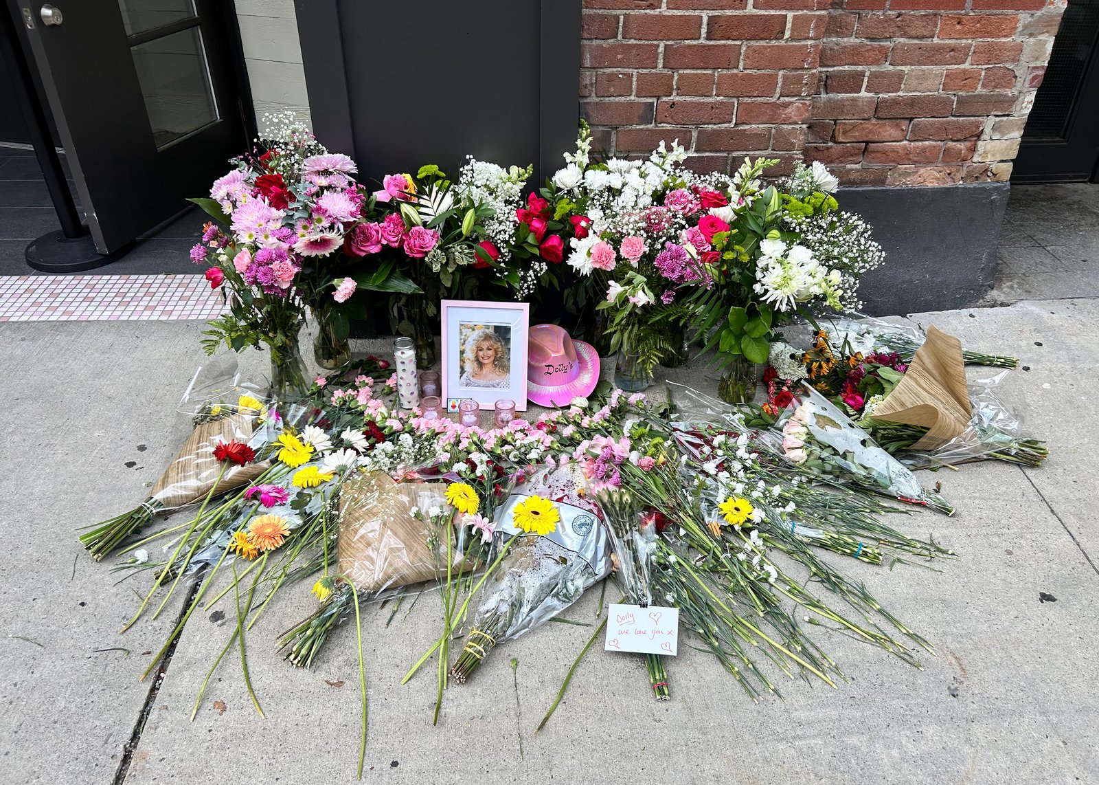 A sidewalk memorial of flowers, candles, a framed photo of Dolly Parton, and a pink Dolly hat left outside Dolly's bar in Toronto after her death