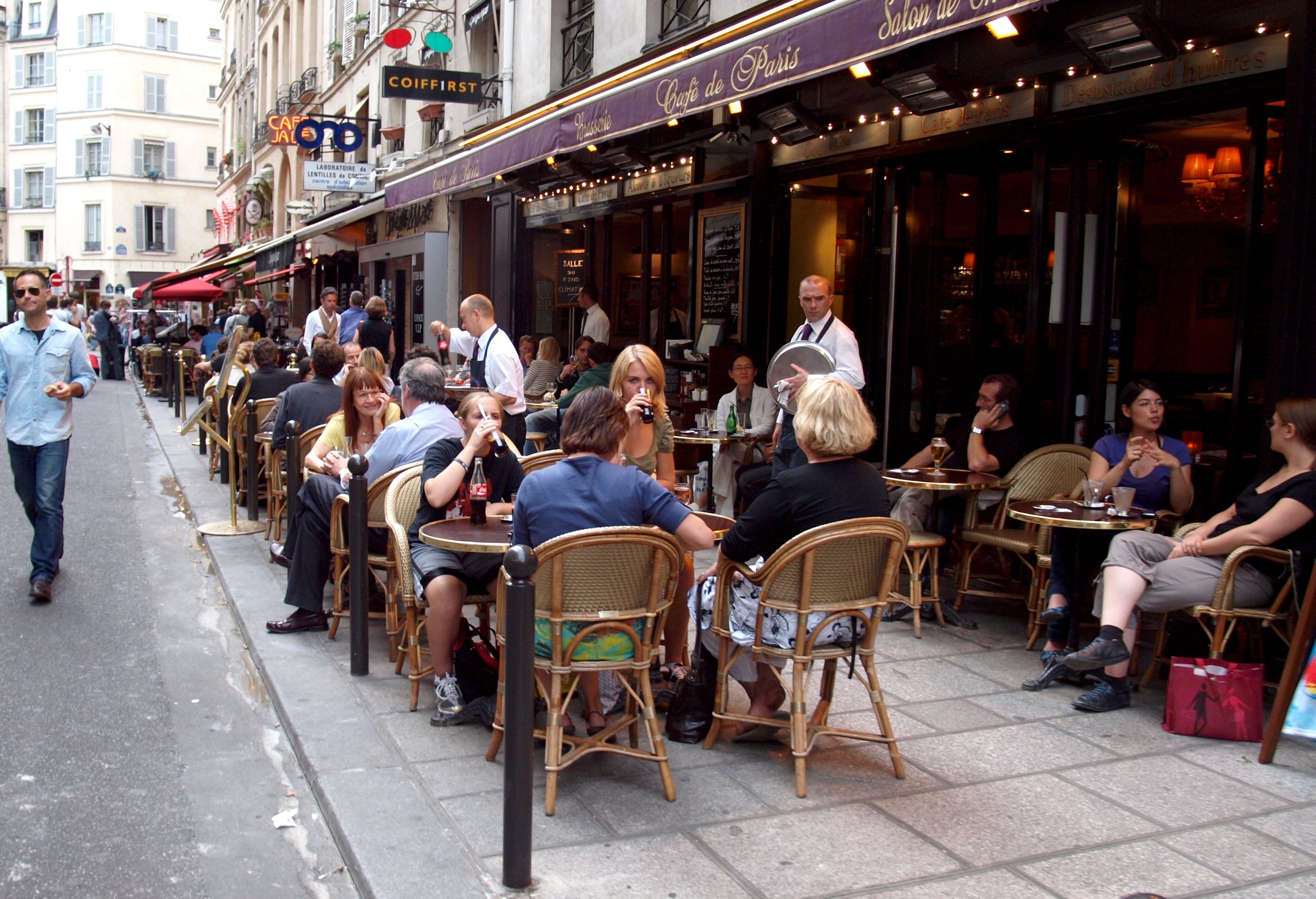 A bustling Parisian café terrace with diners at rattan chairs on a sunny afternoon