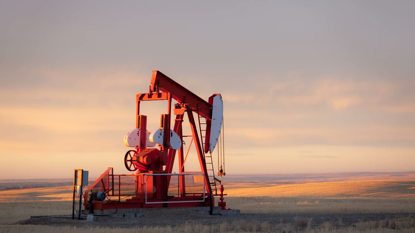 A red oil pumpjack working in a field at sunset, glowing gold against a wide evening sky