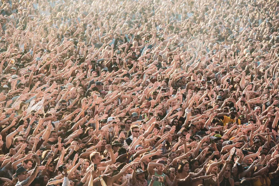 A daytime festival crowd with hands raised in bright sunlight