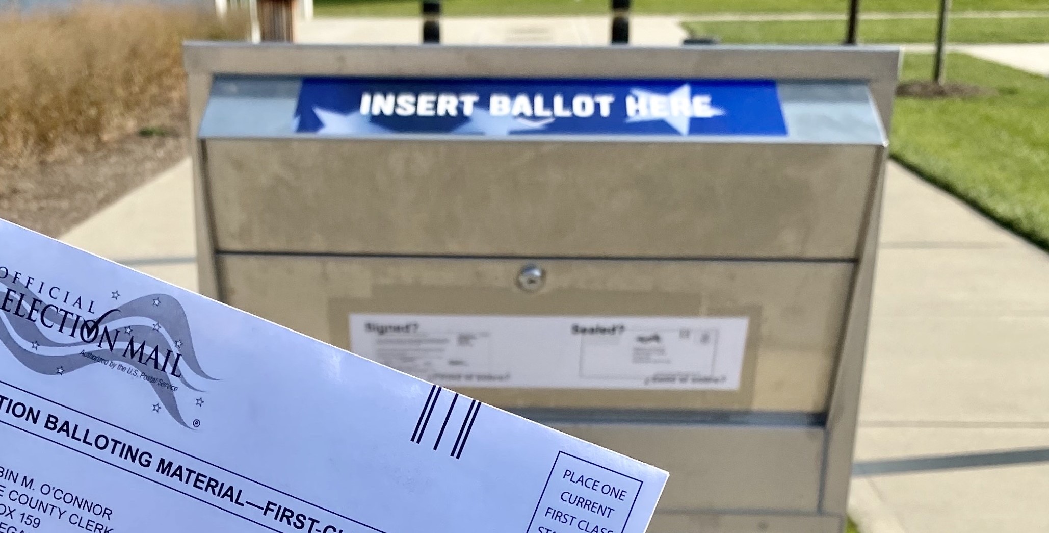A mail-in ballot envelope in front of an official ballot drop box