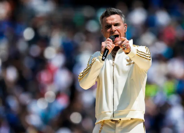 Robbie Williams performing in daylight in a white and gold outfit in 2026.