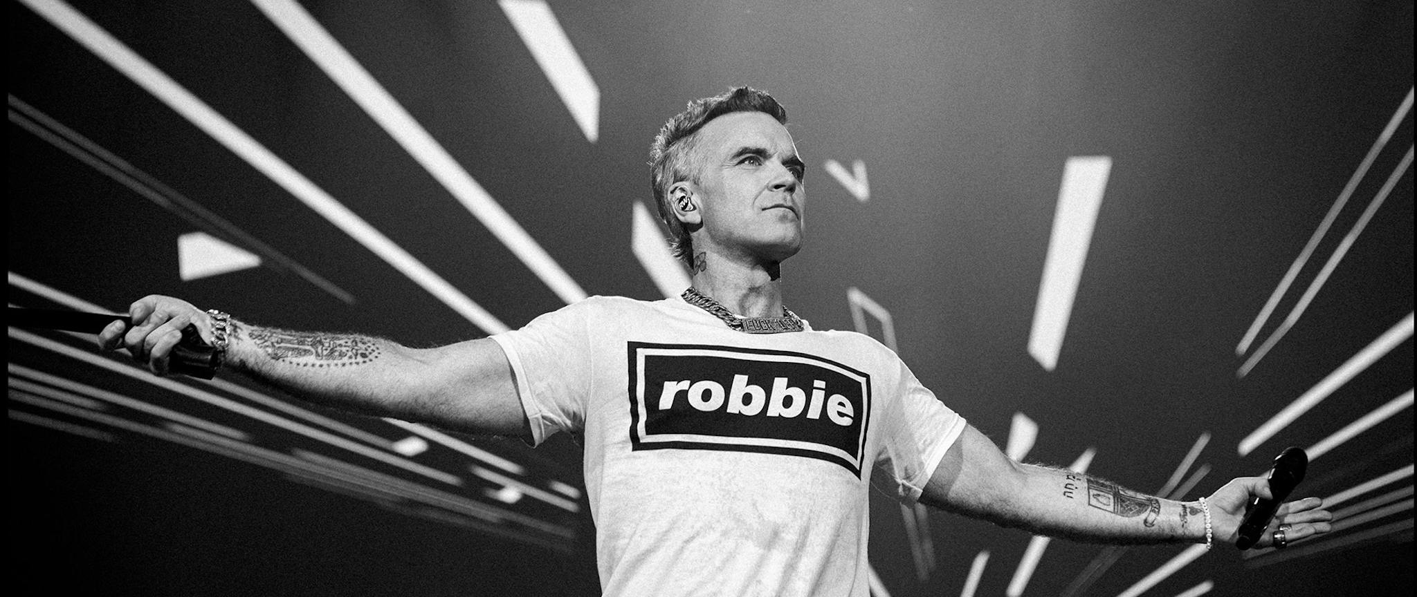 Robbie Williams performing on stage, arms outstretched during his 2026 tour.