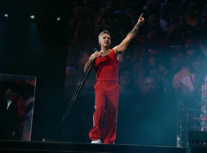 Robbie Williams performing on stage in a red outfit, arm raised to the crowd.
