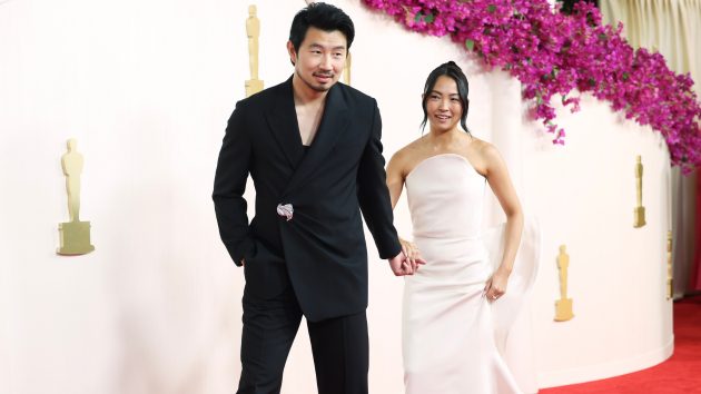 Simu Liu and Allison Hsu walk the red carpet hand in hand at the 2024 Academy Awards, him in black and her in a white one-shoulder gown.