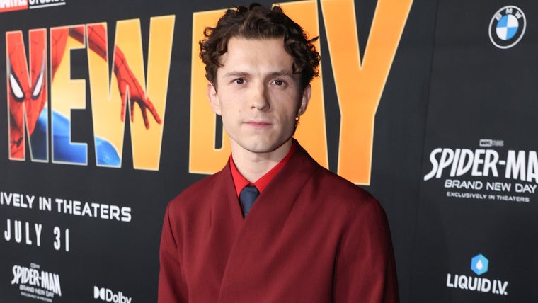 Tom Holland in a burgundy suit at the Spider-Man: Brand New Day premiere, standing in front of a backdrop reading New Day