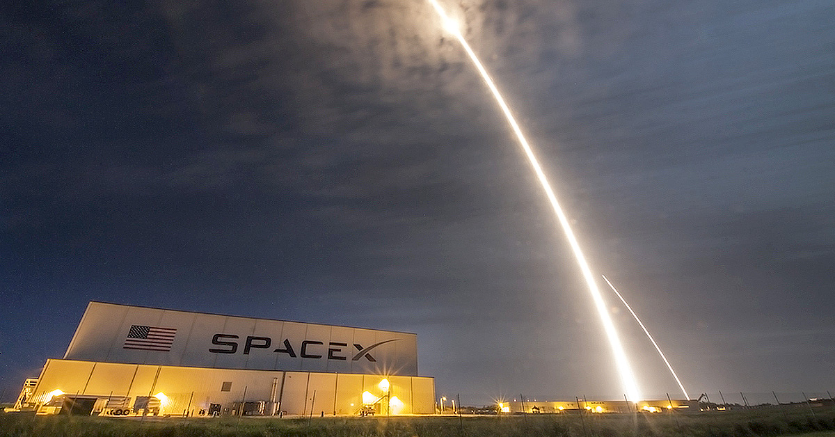 A SpaceX rocket&rsquo;s bright launch trail arcing over a SpaceX facility at night