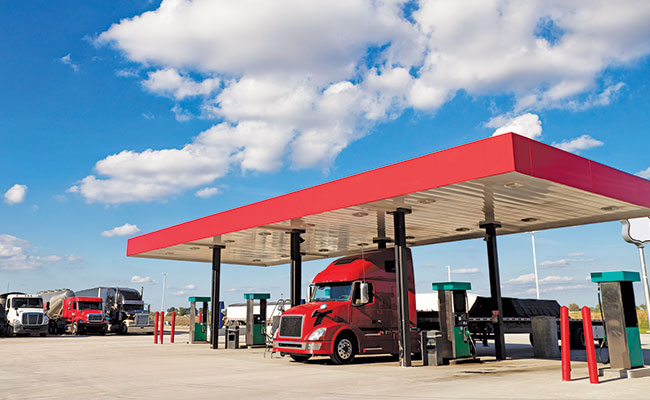 A red semi truck fueling at a truck stop under a blue sky.