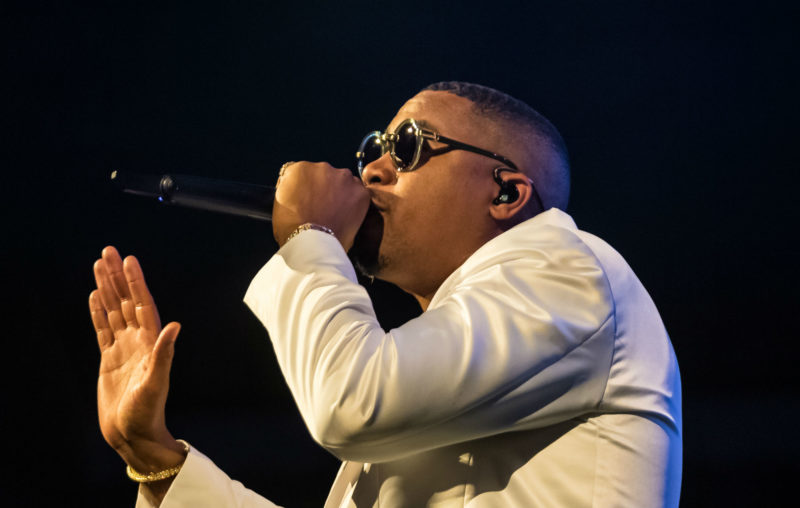 Nas performing live in a white suit and sunglasses, mic in hand